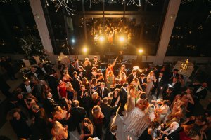 Large group of people in formal attire dance and socialize at an indoor evening event with bright lights and large windows in the background.